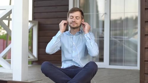 Young man sitting on terrace, listens relaxing music through headphones and meditating