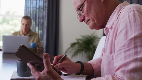 Senior Man Uses Tablet at Home, Woman Works