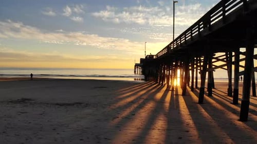 Drone flying under Newport Beach pier at sunset as sunlight beams and people watch the ocean in silh