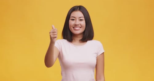 Happy Asian Lady in White T-shirt Gesturing Thumb Up and Smiling at Camera, Orange Studio Background