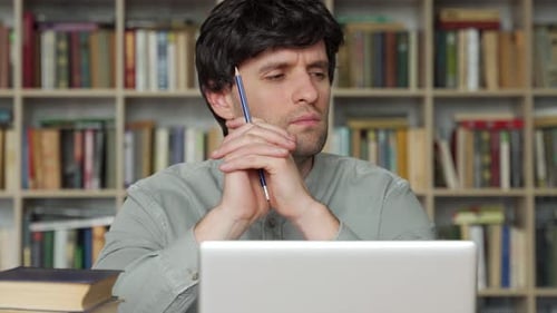 Thoughtful Young Man is Sitting at a Desk in the Office Holding a Pencil in His Hand Looking Away