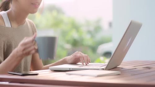 Freelance Asian woman working on laptop and drinking coffee sitting on table in the garden.