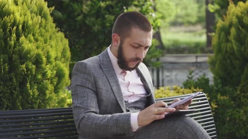 Man Writing on Bench with Tablet in City Park