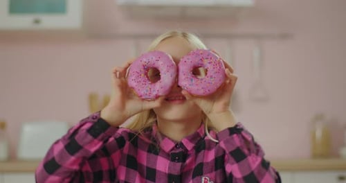Cute Blonde Little Girl in Pink Shirt Holding Two Pink Donuts in Hands By the Face. Pink Donuts Eyes