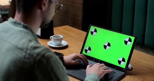 Young Man in Glases Working on Laptop with Green Screen in Coffee Shop