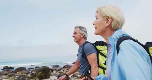 Senior hiker couple with backpack and hiking poles walking while hiking on the beach.