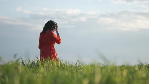 Beautiful Young Woman in a Red Dress Walks in a Green Wheat Field at Sunset or Dawn