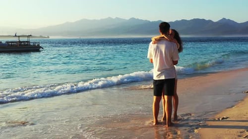 Young couple in love hugging and kissing on exotic beach washed by sea waves with anchored boats on