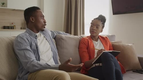 Psycholog Woman Listening to a Young Adult Man and Taking Notes in an Apartment