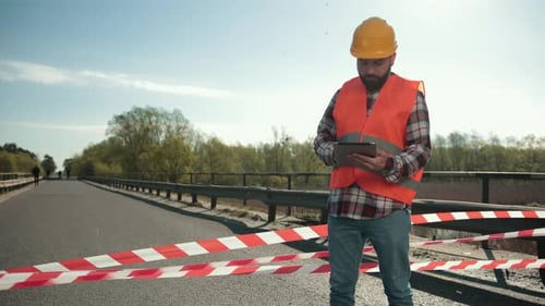 Young Bearded Man Inspector in an Orange Vest and Protective Helmet with Tablet