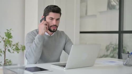 Angry Businessman Talking on Smartphone While Using Laptop in Office