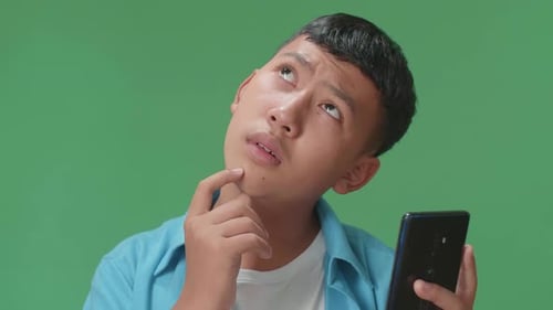 Asian Boy Use Mobile Phone And Thinking About Something While Standing On Green Screen In The Studio