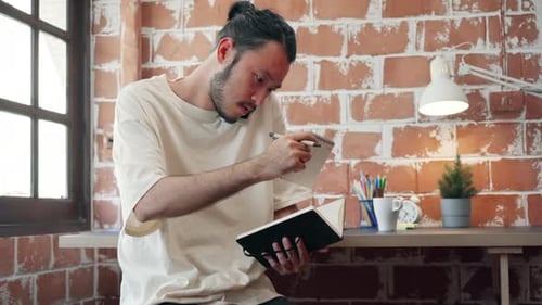 Asiatischer Typ, der an seinem Schreibtisch im Büro arbeitet und einen Anruf auf seinem Handy entgegennimmt, während er Notizen schreibt.