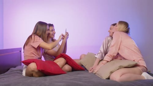Quatro moças bonitas e felizes de pijama sentam-se na cama e tiram selfies no telefone na despedida de solteira
