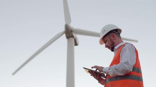 Ein männlicher Ingenieur in einer orangefarbenen Weste und einem weißen Schutzhelm vor dem Hintergrund einer Windmühle sieht aus und