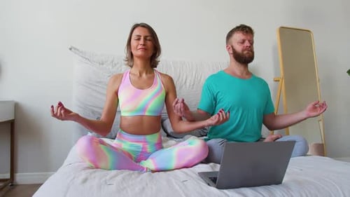 Una pareja de mediana edad medita frente al monitor de un ordenador portátil en una cama bajo la guía de un internauta