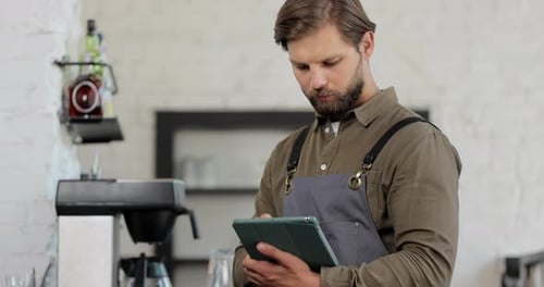 Young Successful Cafe Owner Standing in Cafe Wearing Black Apron Using Digital Tablet While