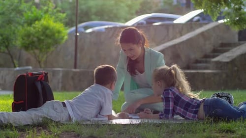 Cheerful Woman Educator Read Book for Little Boy and Girl Sitting on Green Grass in Nature in Sunny