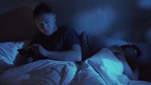 Man Using Phone in Bed While Woman Sleeps