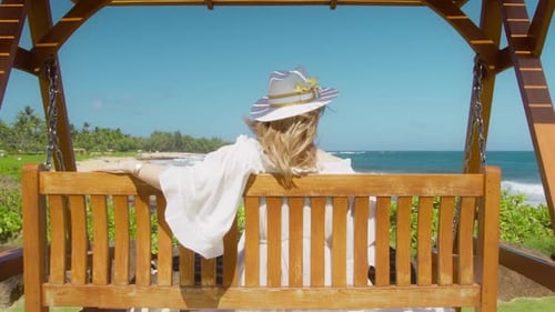 Relaxed Woman in White Hat Swinging with Ocean Breeze Blowing Her Blonde Hair