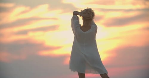 Young Woman with Blonde Hair Wearing White Dress is Dancing on the Lake Pier