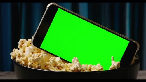 Smart phone with green touch screen and popcorn in a big bowl on the table.