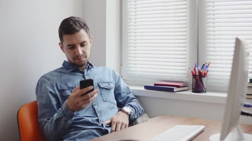 Young Office Worker Using His Phone at the Office Sitting at the Table with Computer Phone and Cup