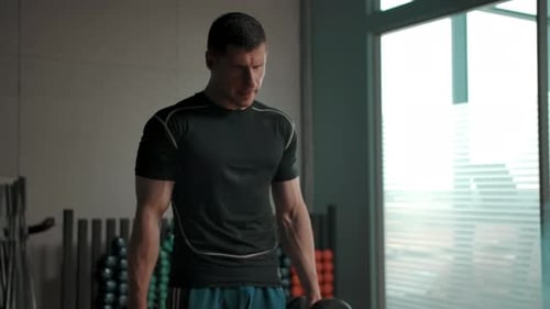 Muscled Man Shakes Biceps with Big Heavy Dumbbells in the Gym Near Huge Windows