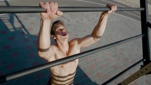 Young Athletic Man is Doing Pull Ups Exercises on Horizontal Bar During Intense Workout Outdoors