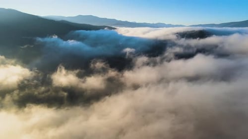Aerial view of landscape above foggy fast moving clouds covering mountain hills at sunrise.