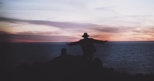 Silhouette eines Mannes, der auf eine Klippe zuläuft und in der Abenddämmerung seine Arme