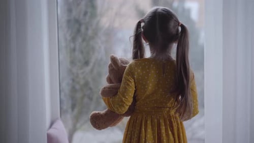Back View of Little Caucasian Girl Hugging Teddy Bear and Looking Out the Window. Brunette Kid