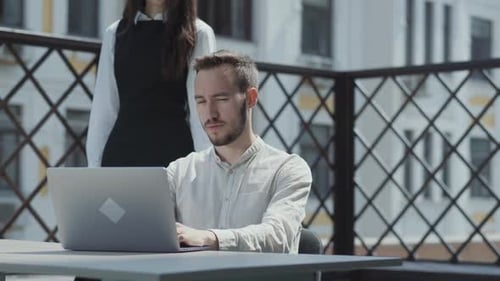 Bärtiger junger Mann sitzt am Tisch auf der Terrasse vor dem Laptop und arbeitet. Hübsches Mädchen