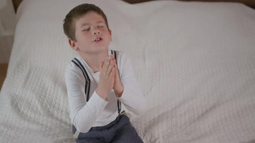 Boy Sits on Bed Praying with Clasped Hands