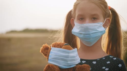 Sad Little Girl and Teddy Bear with Surgical Mask at Sunset. A Child Holding Her Teddy Bear During