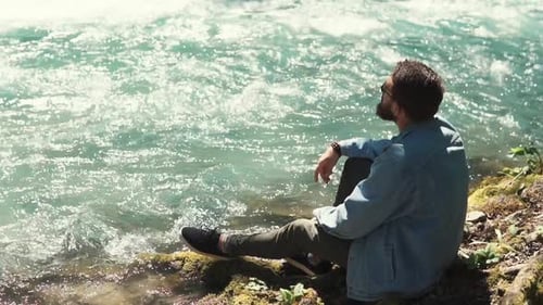 Ein junger Mann sitzt in der Nähe eines schnellen Flusses, er genießt den Wind und die Natur