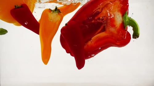 Red and Orange Peppers Fall Into the Water with Splashes and Bubbles, Slow Motion Close-up