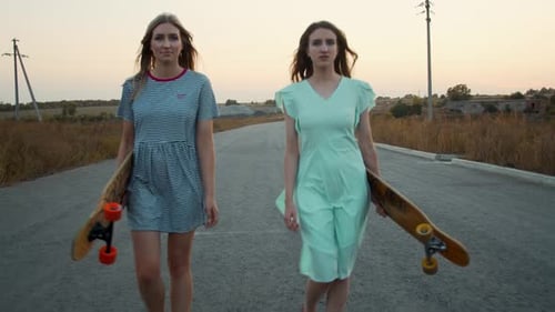 Two Young Women Walk with Skateboards on Road