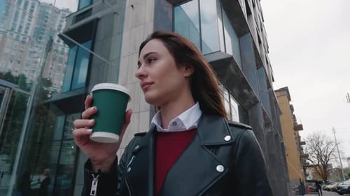 A Confident Businesswoman in a Leather Jacket Walks Down the Street and Drinks Coffee Passing By