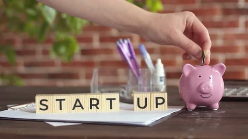 Hand Puts Money in Piggy Bank Next to Which There are Cubes with the Word Start Up