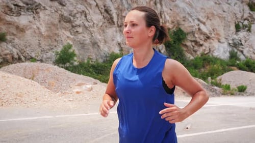 Young smiling motivated woman running on road along rocky cliffs in morning