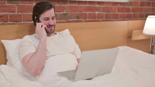 Man Chatting on Phone in Bed With Laptop