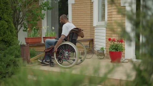 Side View of Paralyzed African American Man in Wheelchair Smelling Flowers in Pot on Backyard Porch