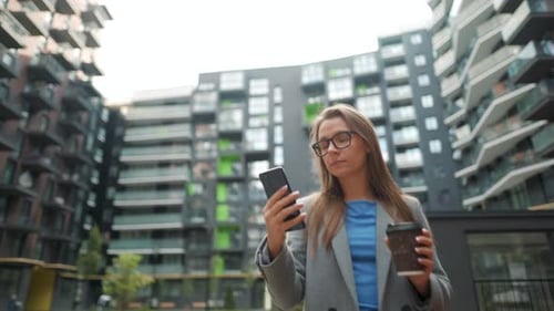 Formally Dressed Woman Walking Down the Street in a Business District with Coffee in Hand and Using