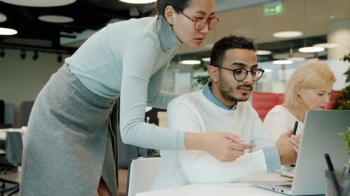Asian Woman and Arab Man Talking Looking at Laptop Screen in Shared Workspace Office