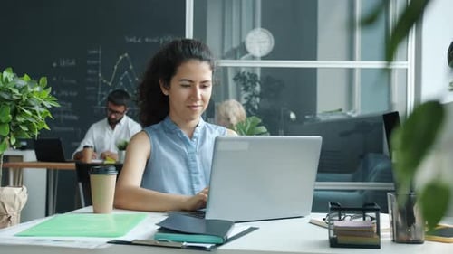 Cheerful Young Woman Using Laptop Typing While Colleagues are Working in Background in Shared Office