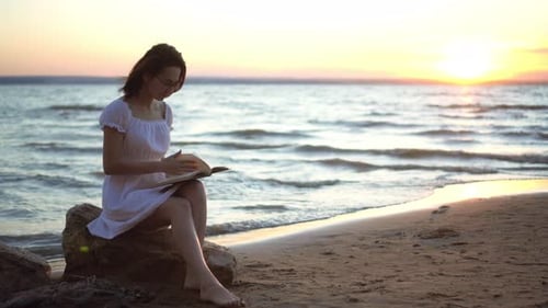 Una mujer joven se sienta en una piedra en la playa junto al mar con un libro en las manos. Una chica vestida de blanco