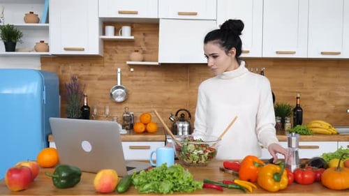 Woman Makes Salad While Using Laptop in Kitchen