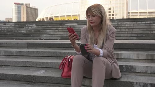 A woman is sitting on the stairs in the center of a big city listening to music.