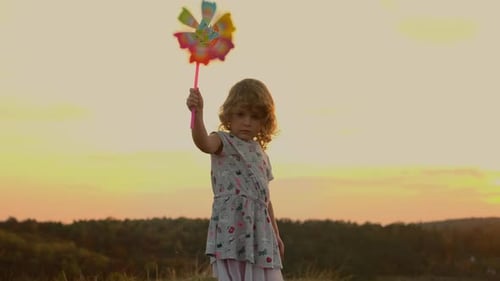 Little Girl Standing On Nature At Sunset. Girl Holding Toy Windmill Moving In The Wind.
